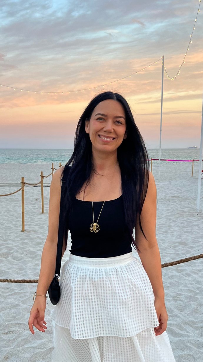 Hypnotherapy Miami - A woman with long black hair smiling at the beach during sunset. She is wearing a black top, a white skirt, and a necklace with a crab pendant. String lights are hanging in the background, and a ship is visible on the horizon.